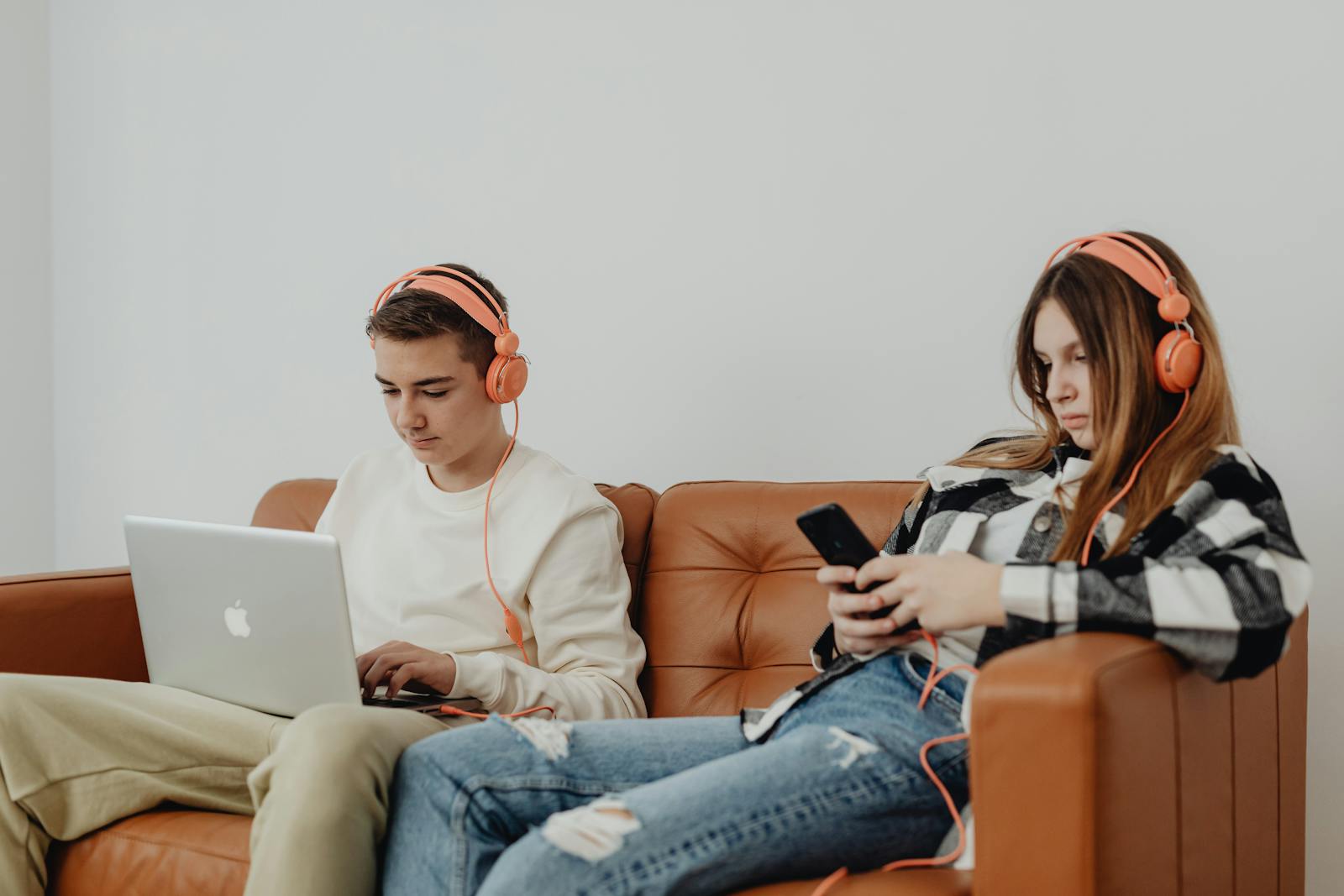 Teens using headphones with laptop and smartphone, lounging on a sofa.